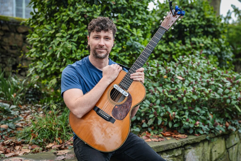 Aodán Coyne seated outdoors holding an acoustic guitar
